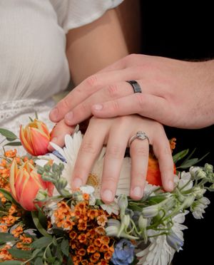 Close-up of hands with wedding rings resting on a colorful bouquet with orange and white flowers.