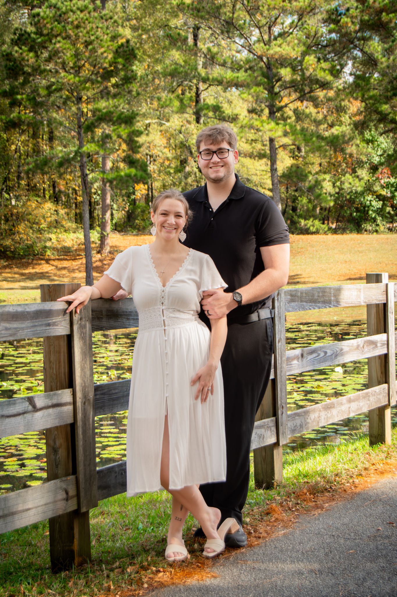 A couple poses together on a wooden bridge overlooking a lily pond surrounded by autumn trees.