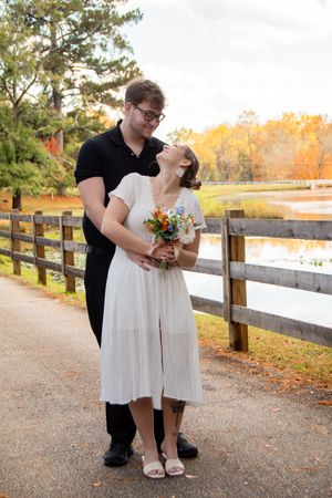 A romantic couple poses together on a gravel path with wooden fencing and autumn trees in the background.