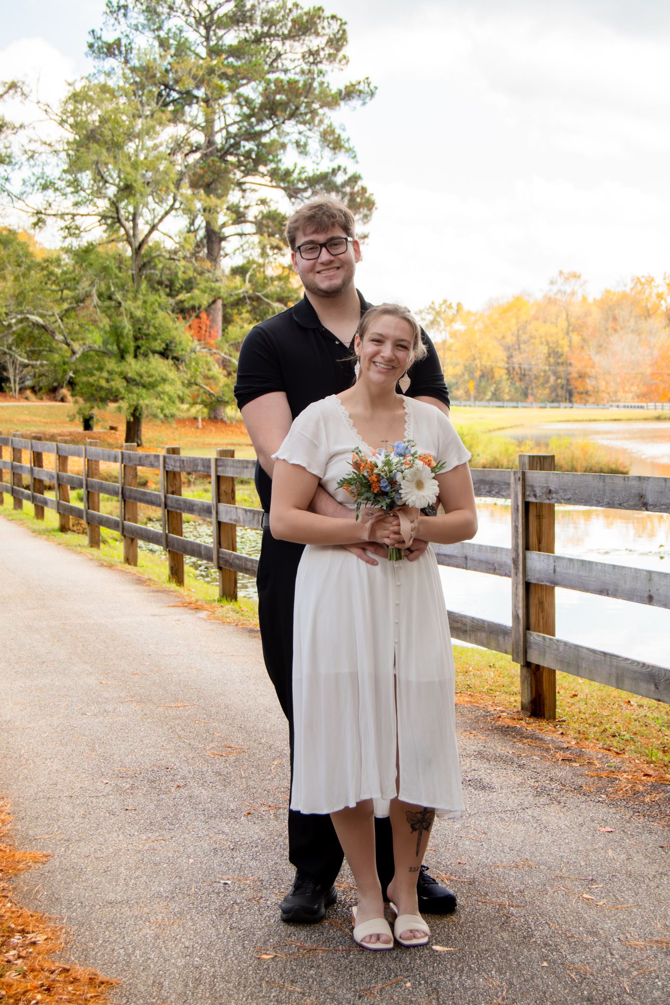 A couple poses together on a wooden bridge path surrounded by autumn trees and fallen leaves.