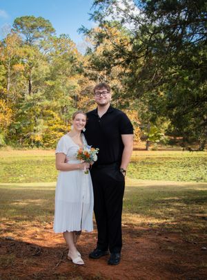 A couple poses together on fallen pine needles with autumn trees creating a natural backdrop for their wedding photos.