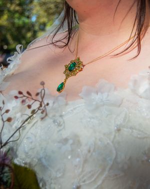 An ornate emerald and gold pendant necklace rests against white lace wedding dress fabric.