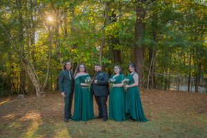 Wedding party members in emerald green dresses pose together in a forest setting during golden hour.