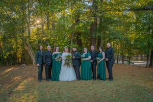 Wedding party poses together in a wooded outdoor setting with green dresses and dark suits.