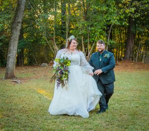 A couple in wedding attire share a playful moment on a grassy lawn surrounded by autumn trees.