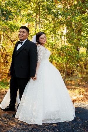 A newlywed couple stands back-to-back in a wooded setting, with golden sunlight filtering through autumn foliage.