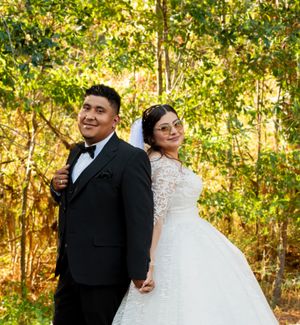 A wedding couple poses together in a sunlit wooded area wearing traditional formal attire.