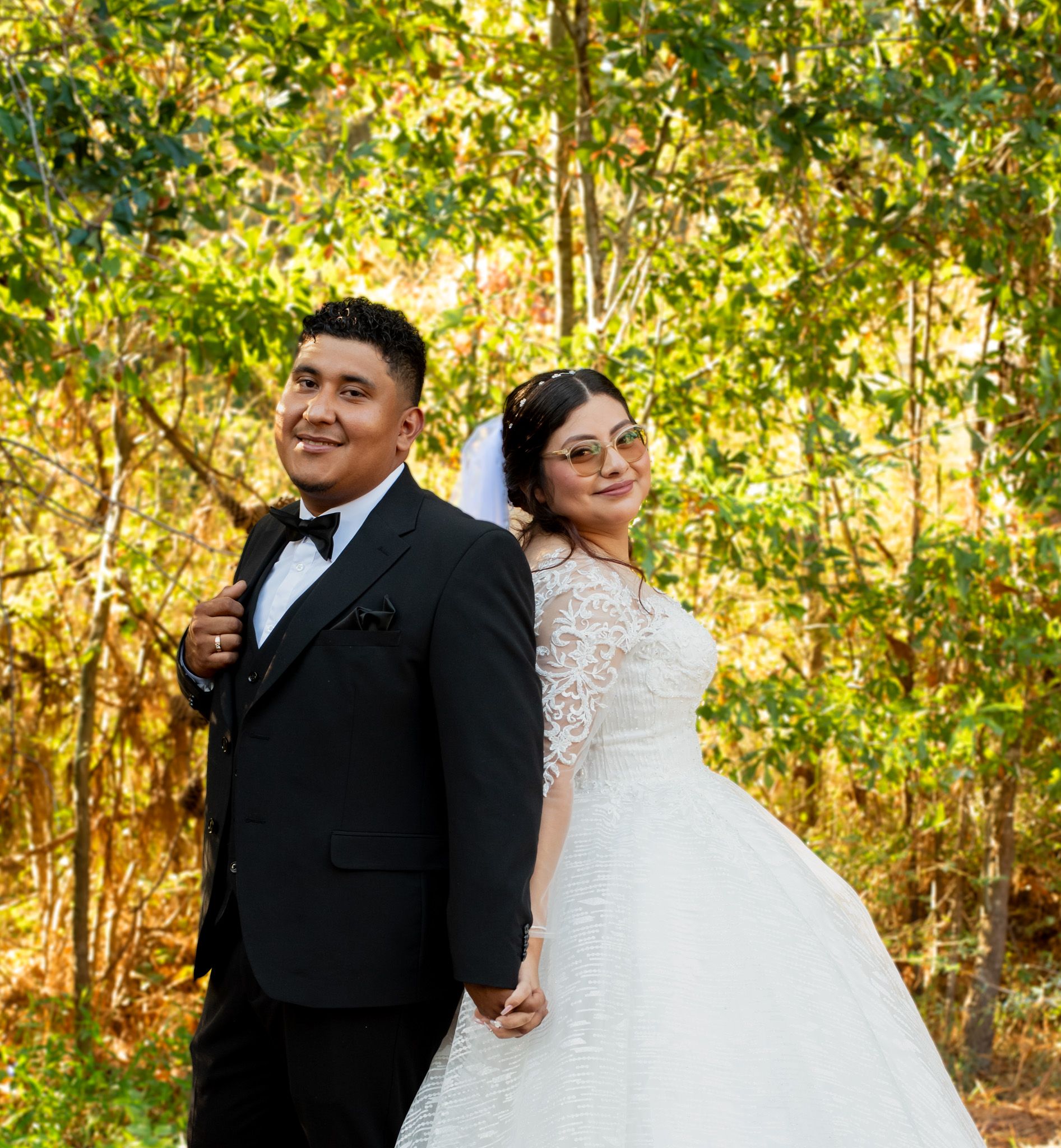 A newlywed couple in formal wedding attire poses together in a sunlit wooded area with golden foliage.