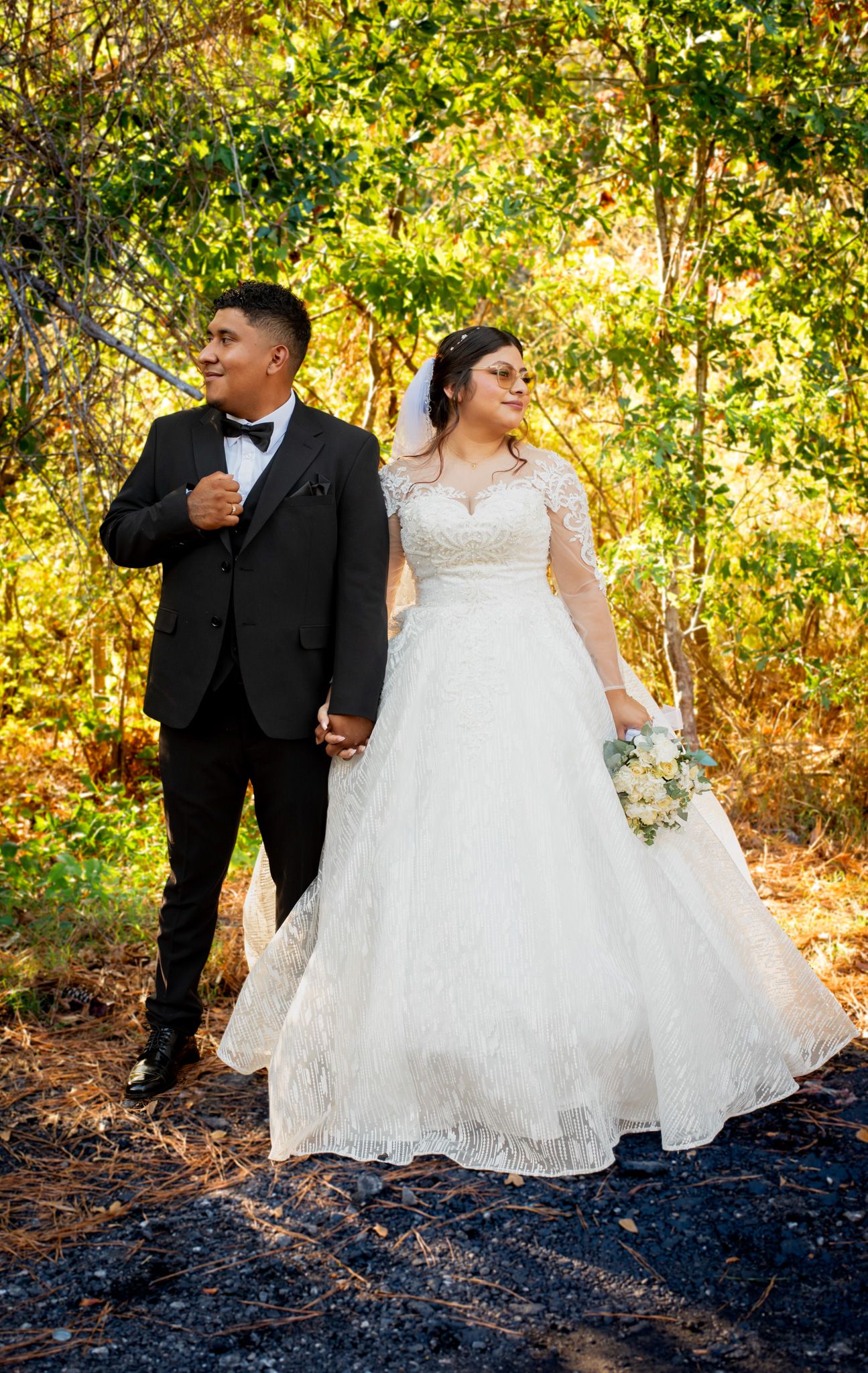 A wedding couple poses in a sunlit autumn forest setting, with the bride in a lace gown and groom in a black tuxedo.