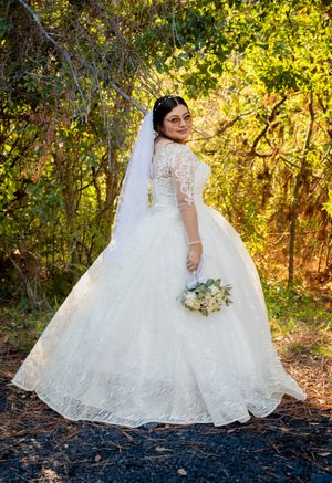 A flowing white wedding dress with intricate lace details photographed in a sunlit wooded setting during golden hour.