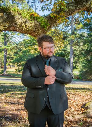 A person in a dark suit and tie stands thoughtfully under a large tree branch in an autumn park setting.