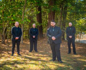 Groomsmen in dark suits stand in line outdoors against a wooded autumn backdrop.