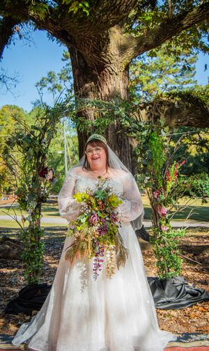 Bride with autumn bouquet poses joyfully under large tree in outdoor wedding setting.