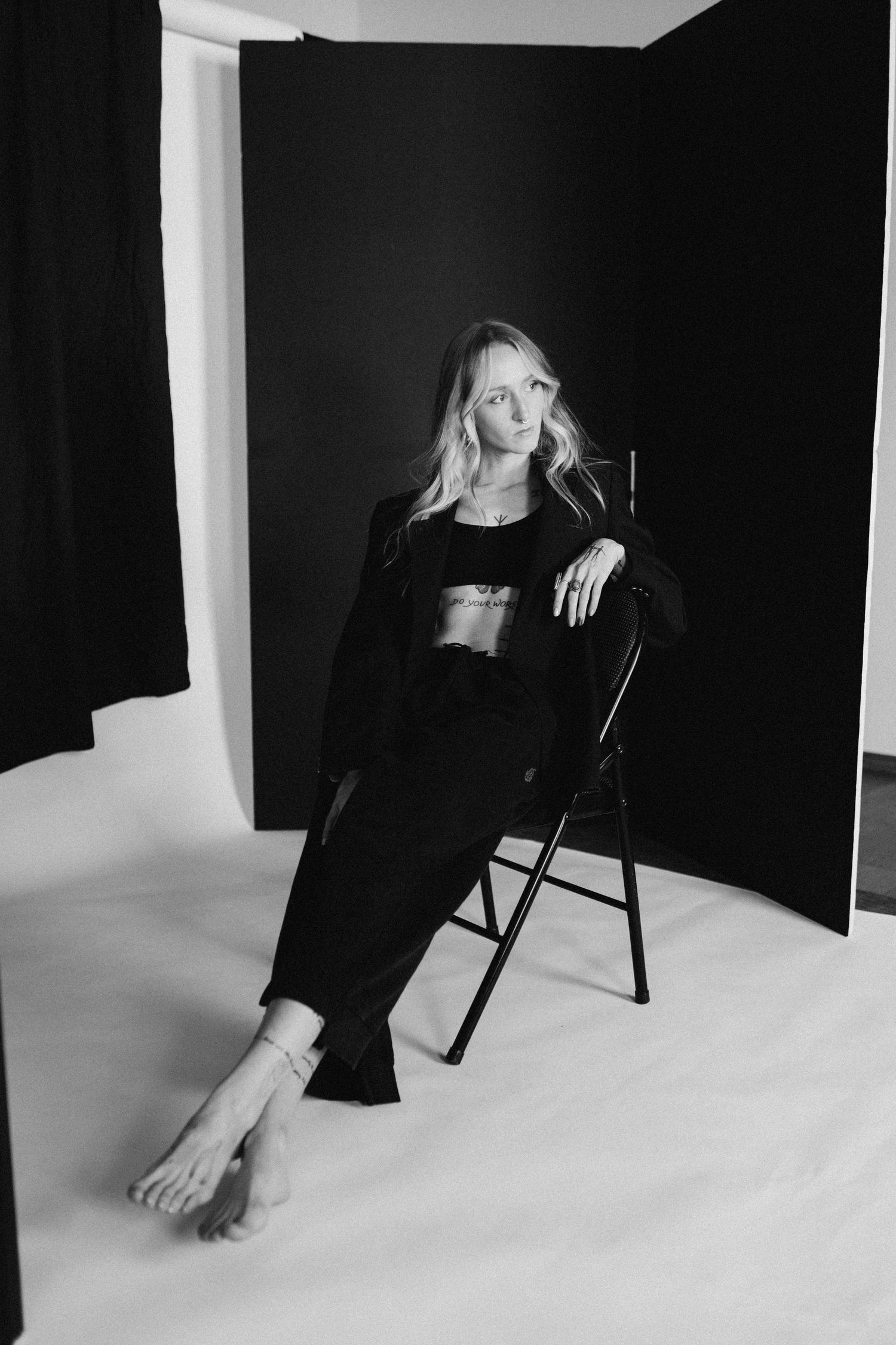 Black and white studio portrait of a blonde woman in all-black outfit seated on a folding chair.