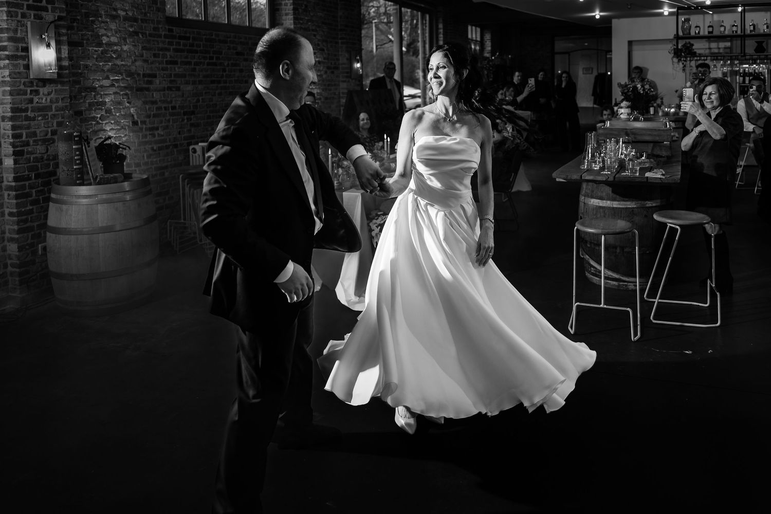 Black and white photo of a couple dancing together in an elegant wedding reception venue with brick walls and a bar.