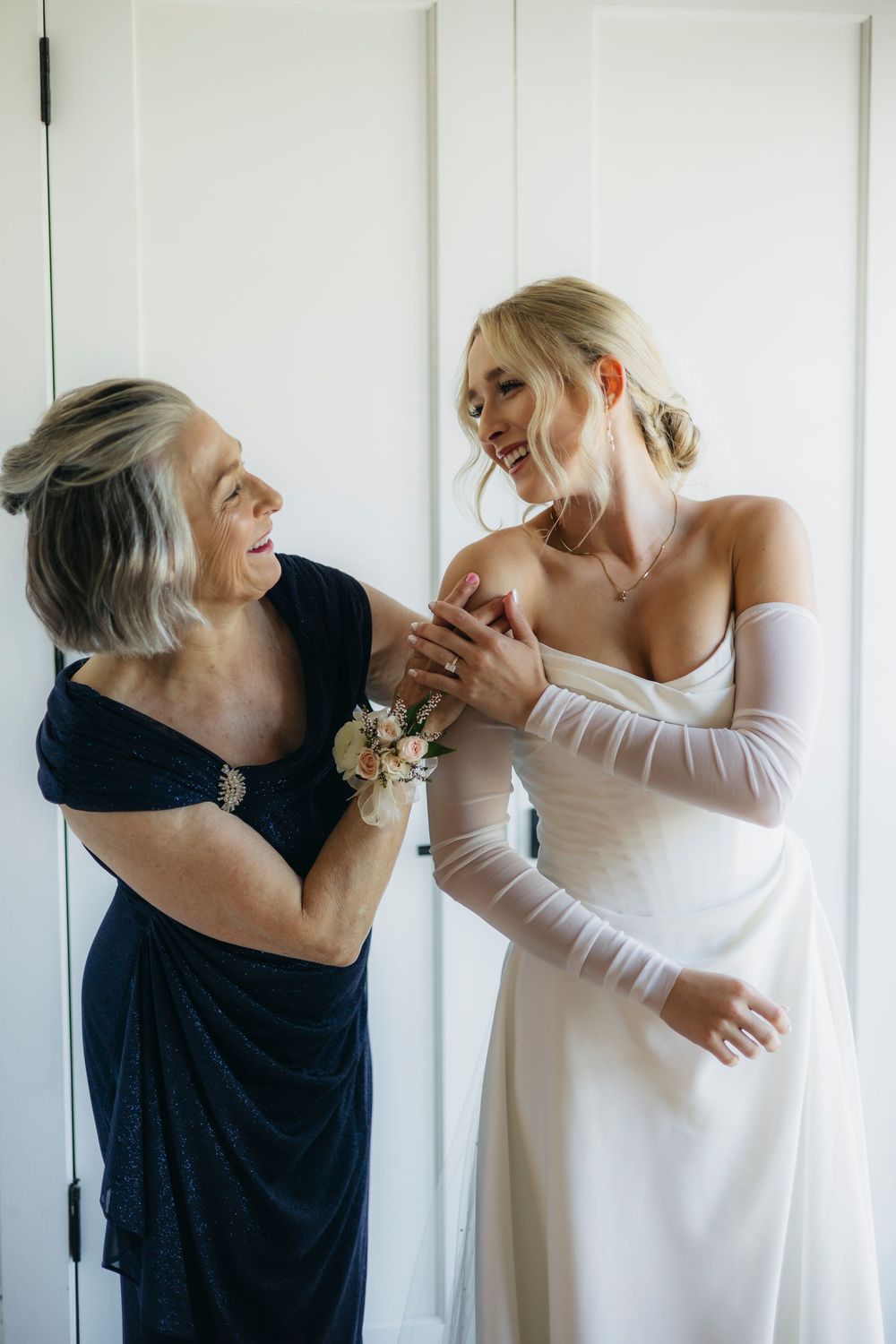 Two people share a tender moment while getting ready, one wearing a navy dress and the other in a white wedding gown.