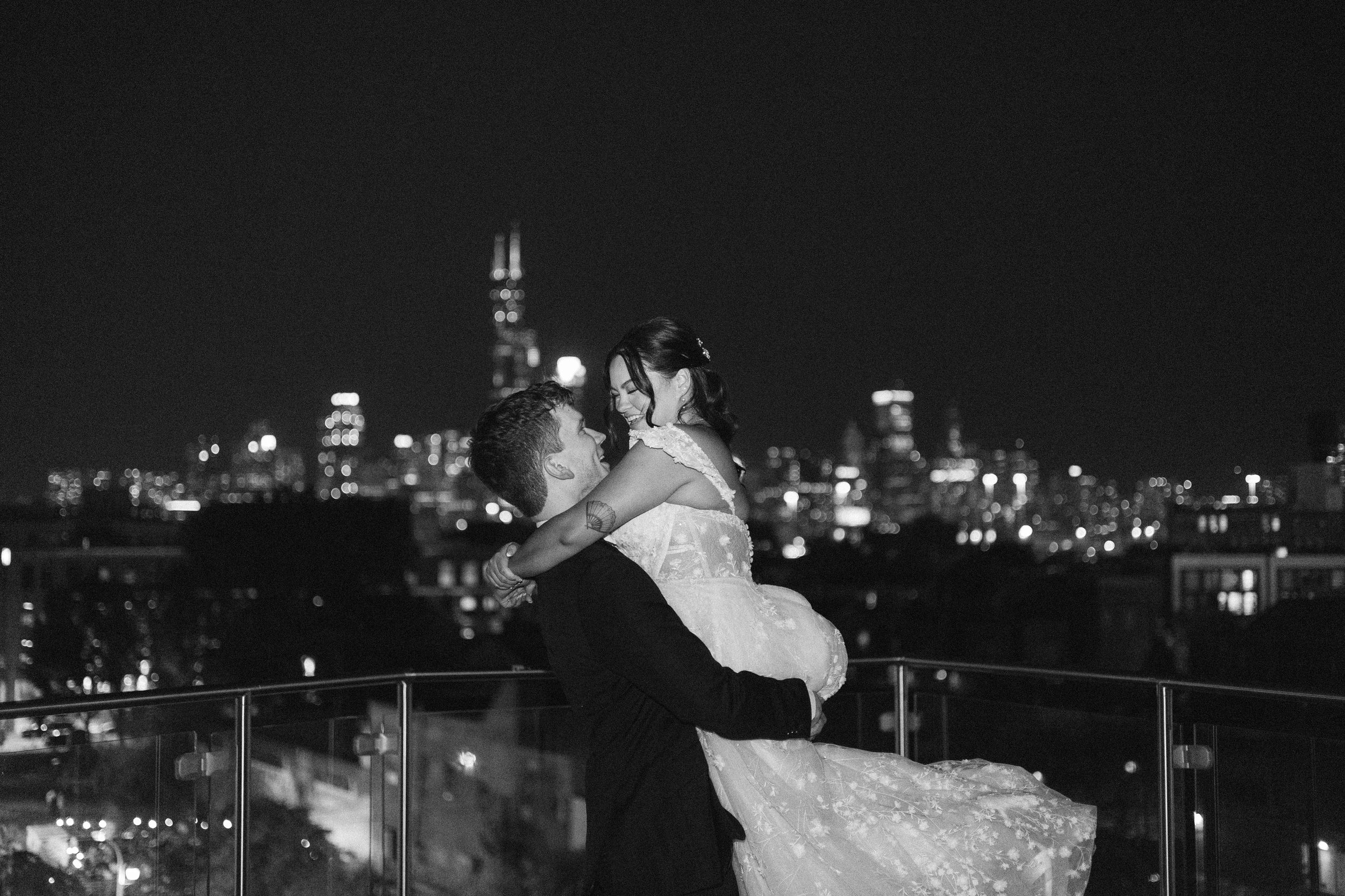 A black and white photo of newlyweds hugging happily on a rooftop with Chicago night skyline on the background