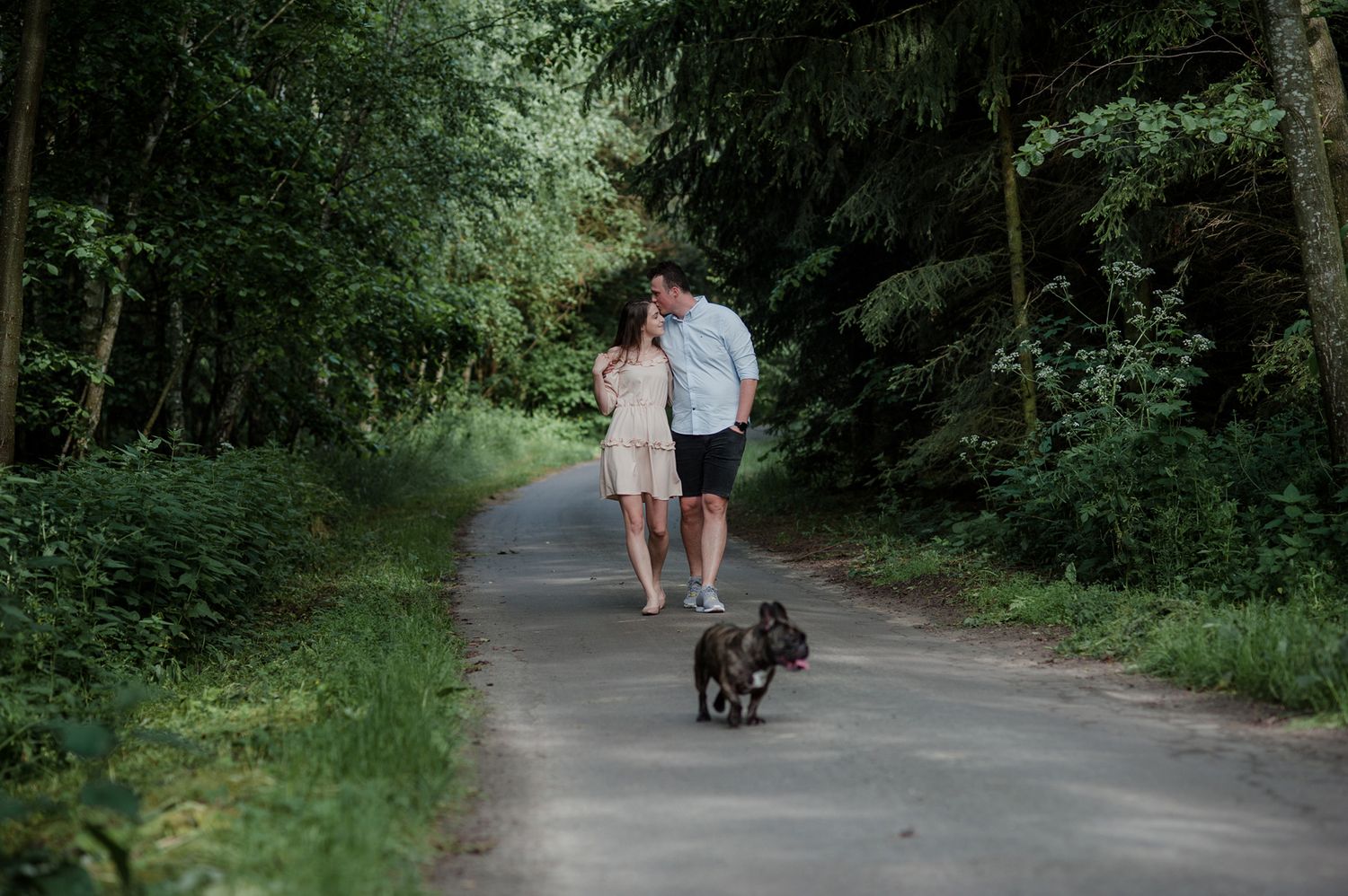 A couple walks their small black dog along a peaceful tree-lined path in a lush green forest.