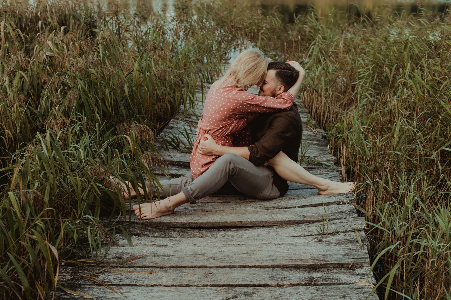 Silhouetted figures share an intimate moment on a rustic wooden walkway at sunset among reeds.