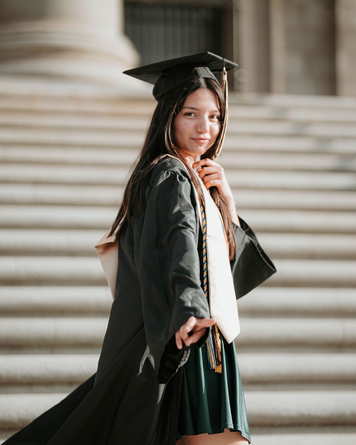 Graduate in black cap and gown poses on stone steps during graduation photoshoot.