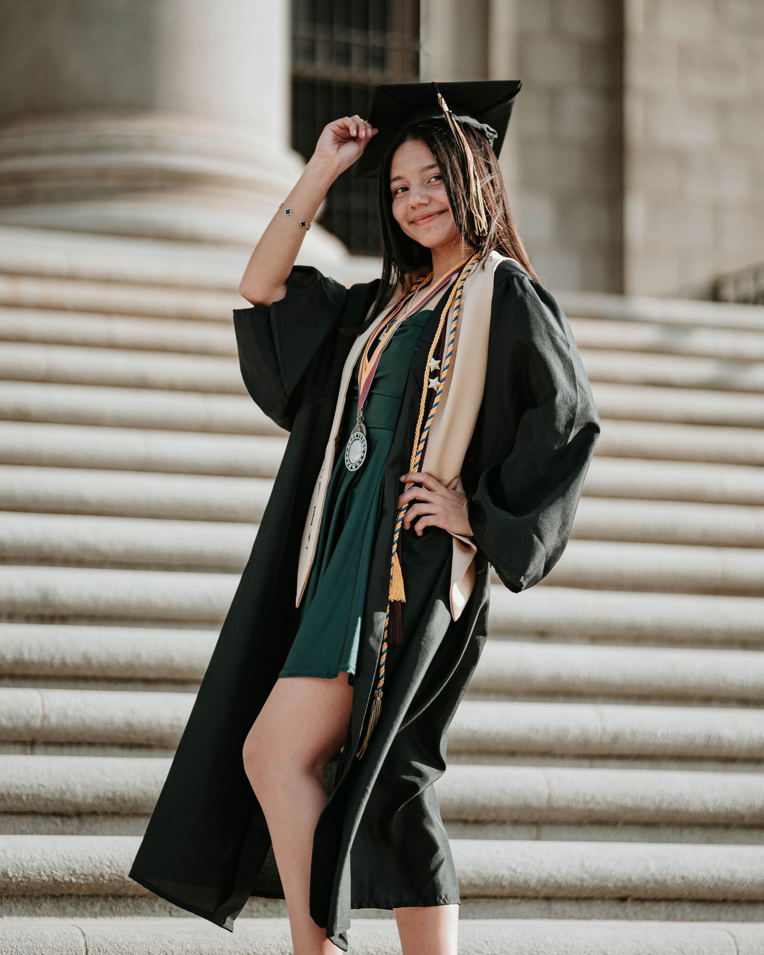 Graduate in academic regalia sits elegantly on concrete steps during outdoor photo session.