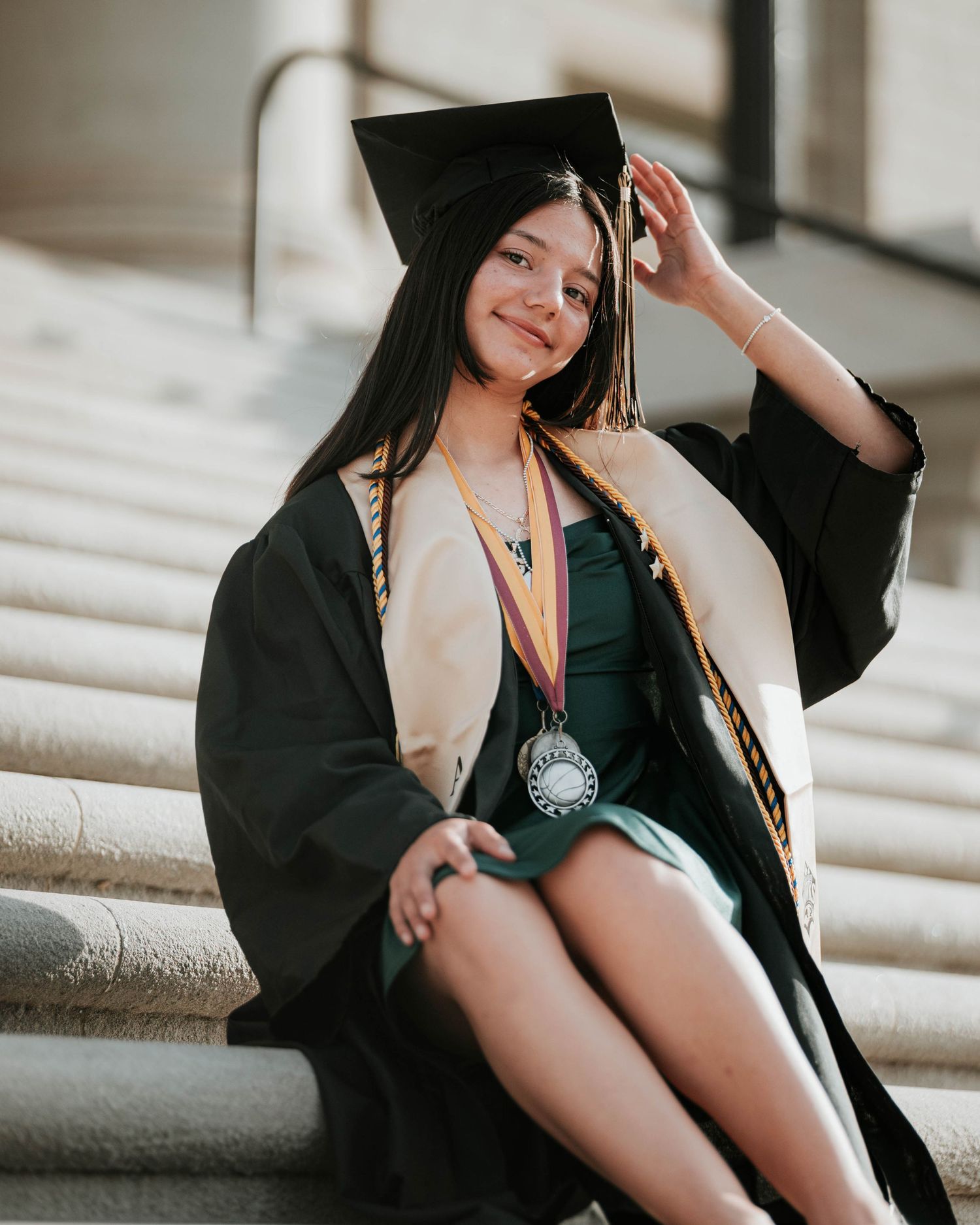 Graduate in black graduation attire poses casually on stone staircase.