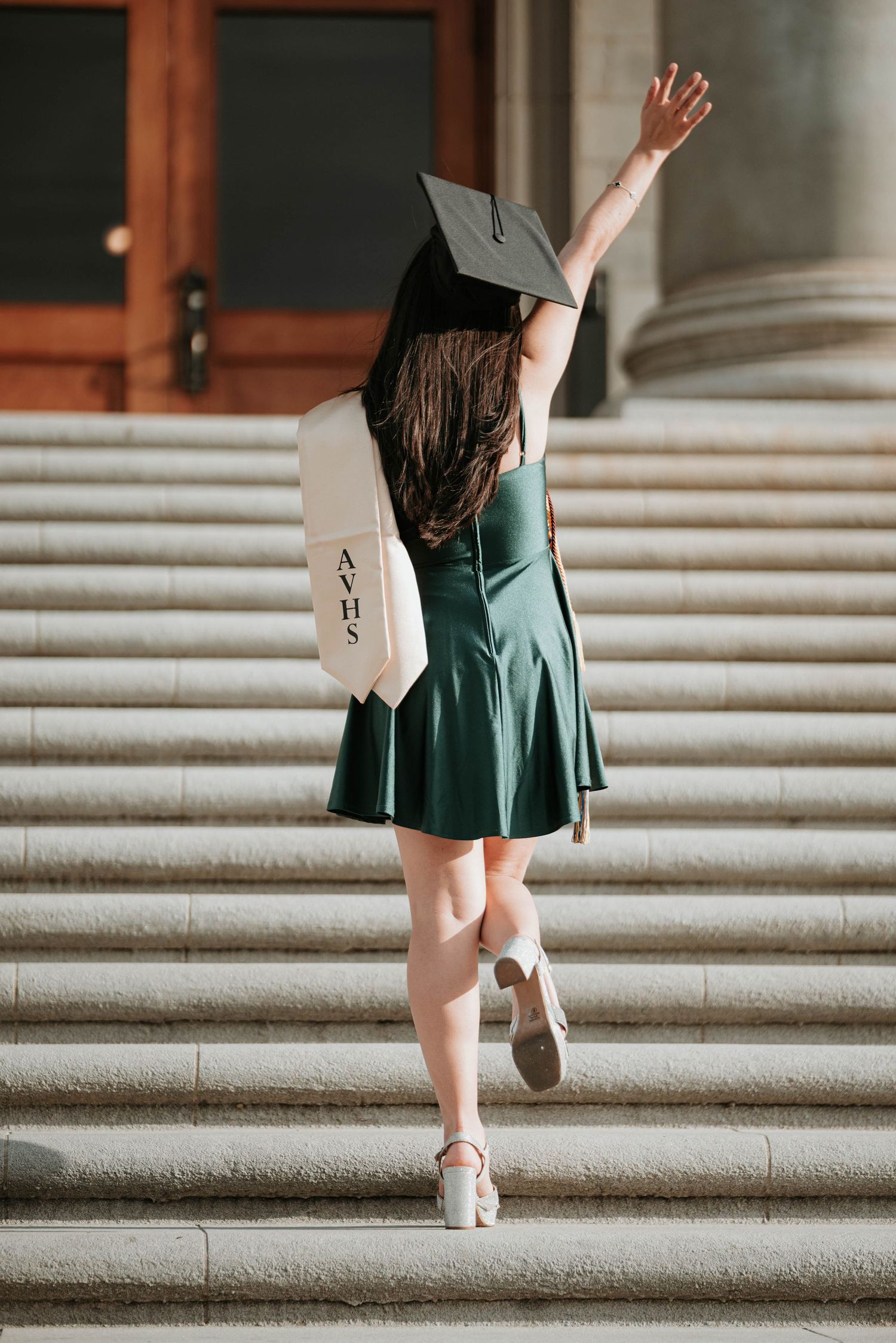 Graduate in emerald green dress and cap ascending stone steps while waving during commencement ceremony.