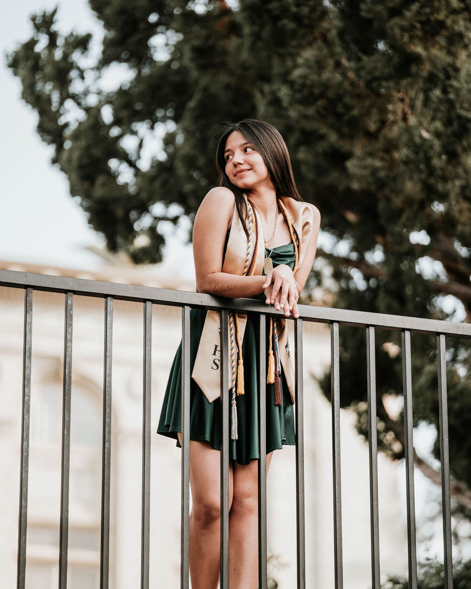 Outdoor portrait session featuring dark dress and white blazer against metal railing with trees in background.