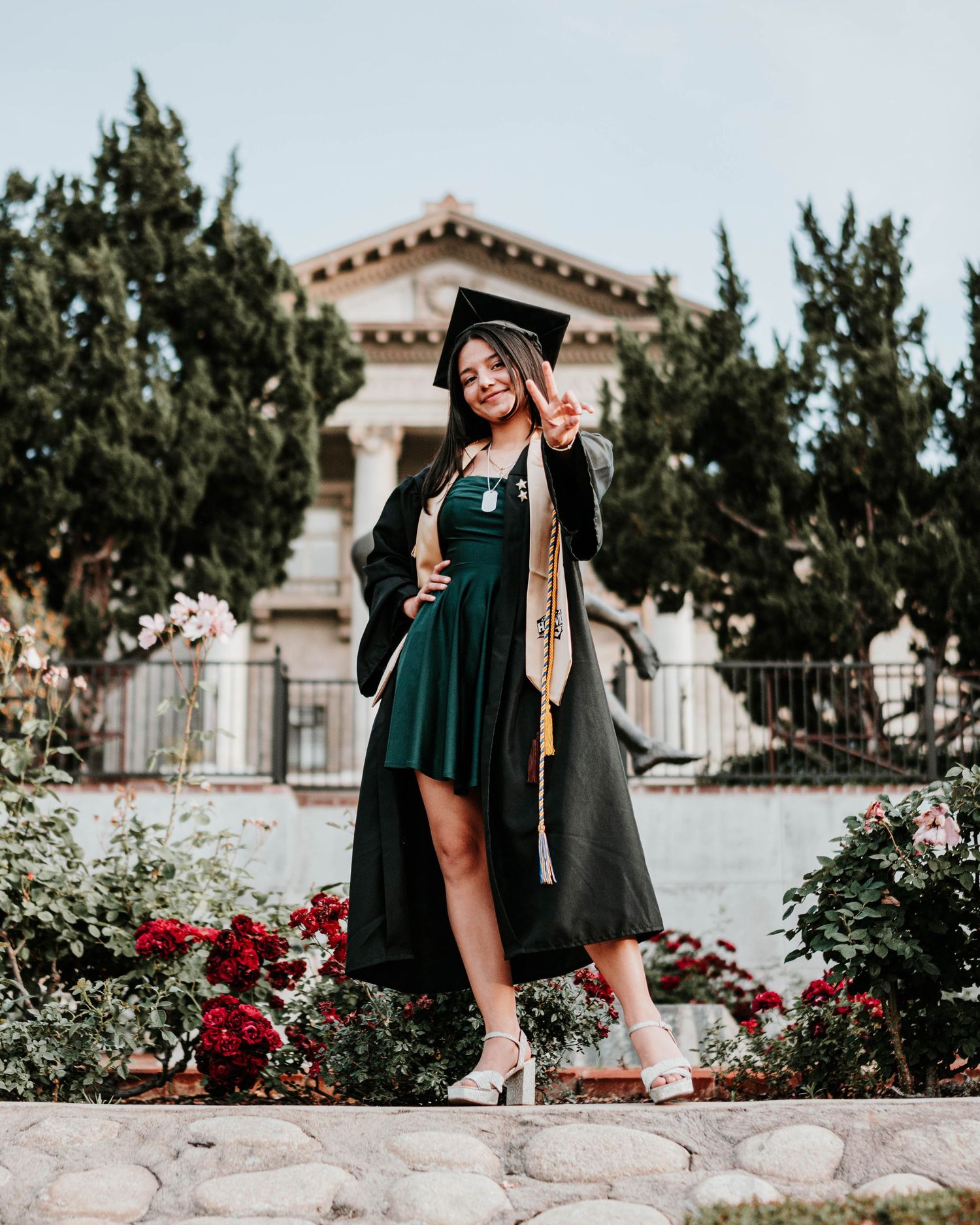 Graduate in emerald green cap and gown poses for graduation photos in a garden setting with red roses and stone walls.