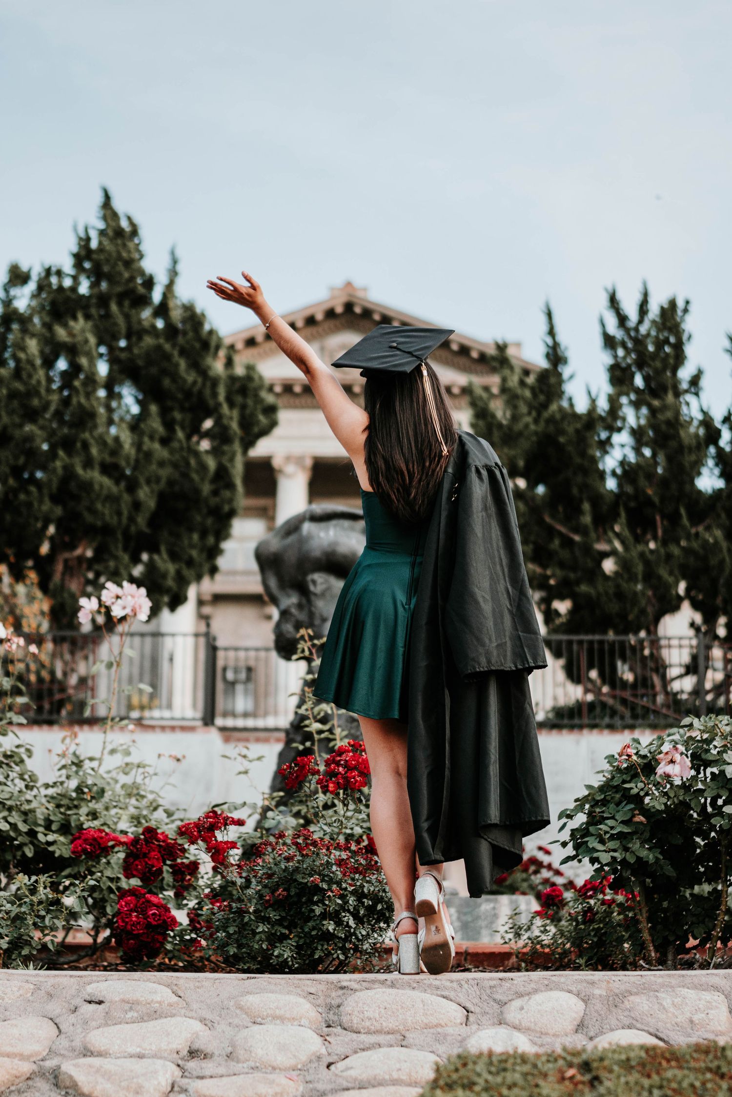 Graduate in cap and gown twirls joyfully in a garden with blooming red roses and stone walls.