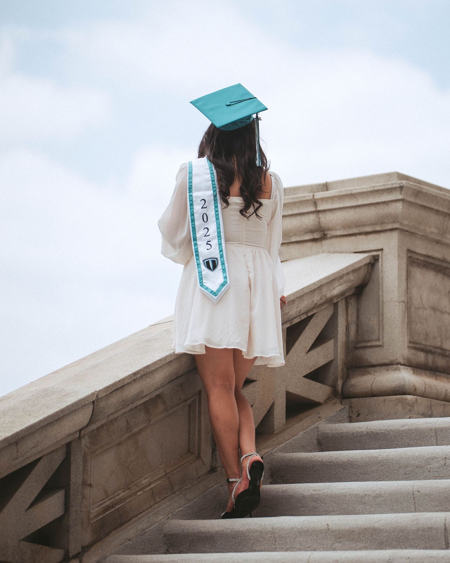 Graduate in white dress and teal cap walking up stone stairs in artistic cropped photo series.