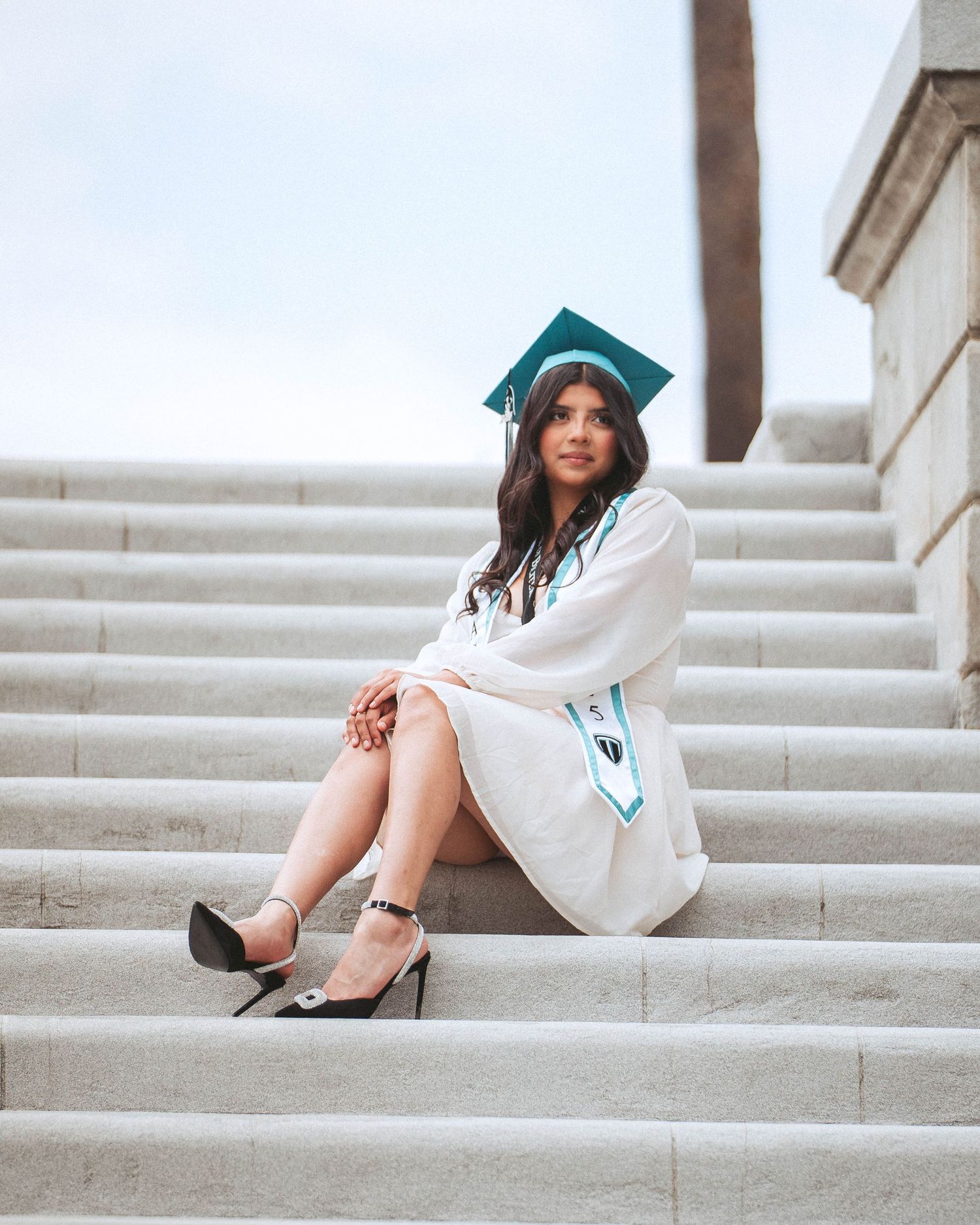 Graduate in white dress and teal cap sits on concrete steps during graduation photo shoot.