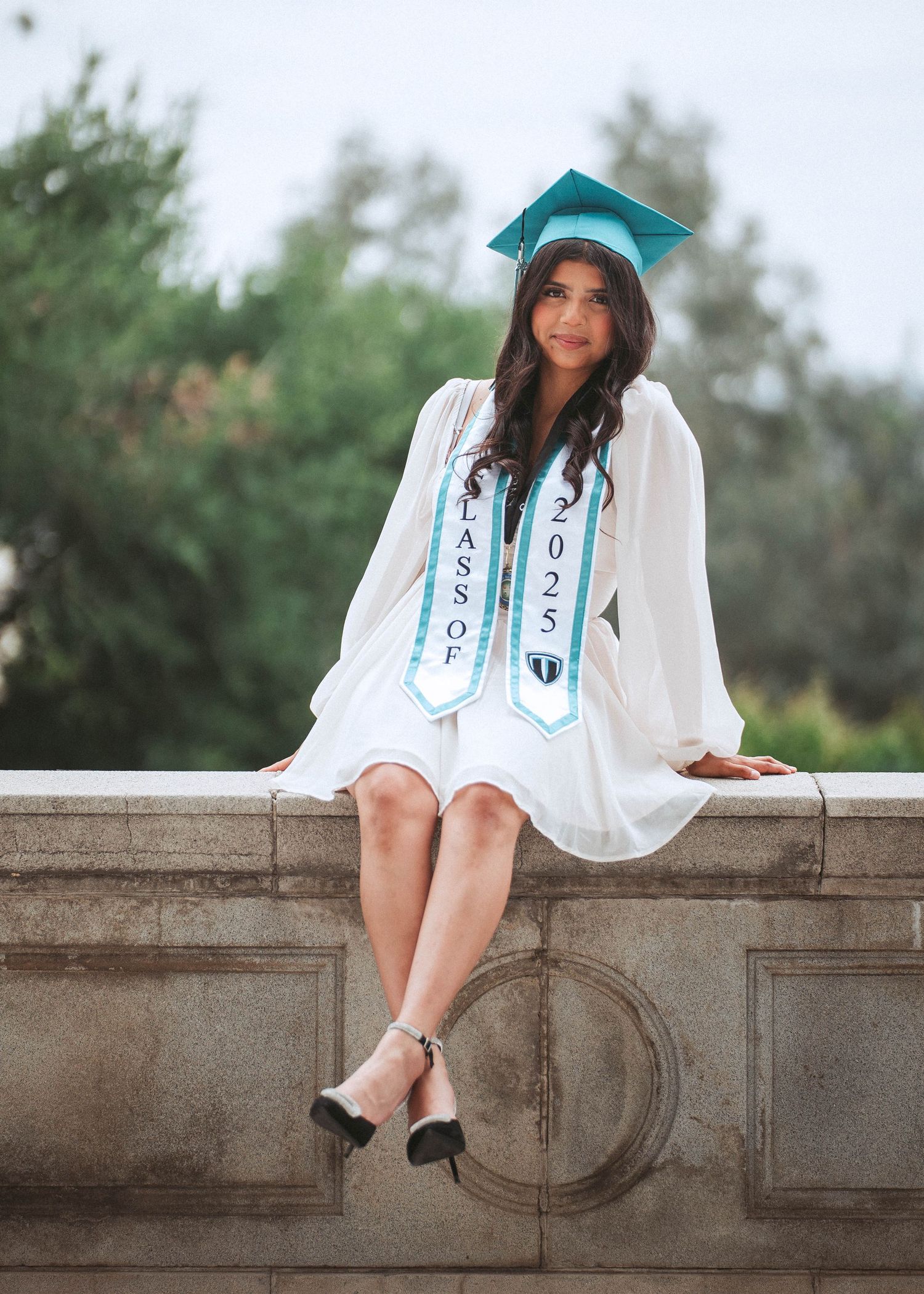 Graduate in white dress and teal cap sits on stone wall during outdoor graduation photoshoot.
