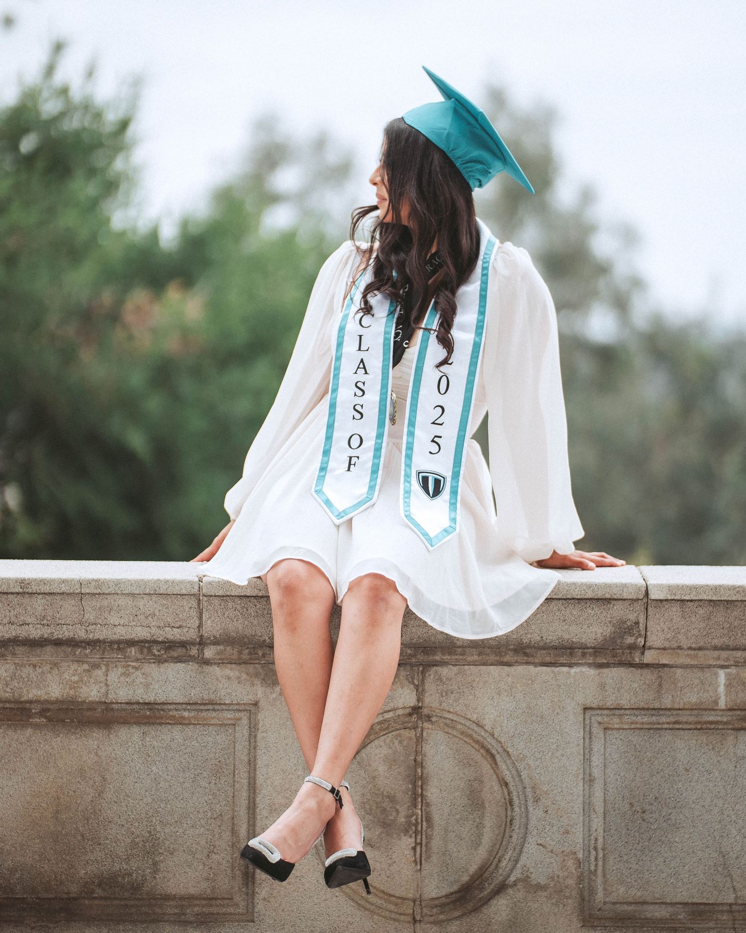 A person in a white dress sits on a stone wall wearing turquoise graduation stoles in an outdoor setting.