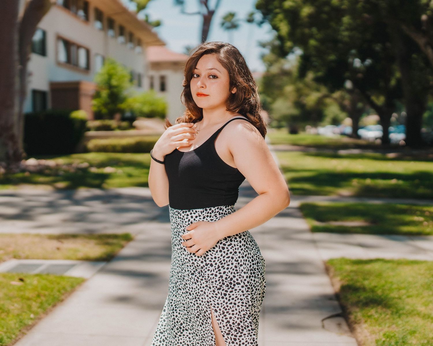 A pregnant woman in a floral dress stands on a sunny sidewalk during a maternity photoshoot.