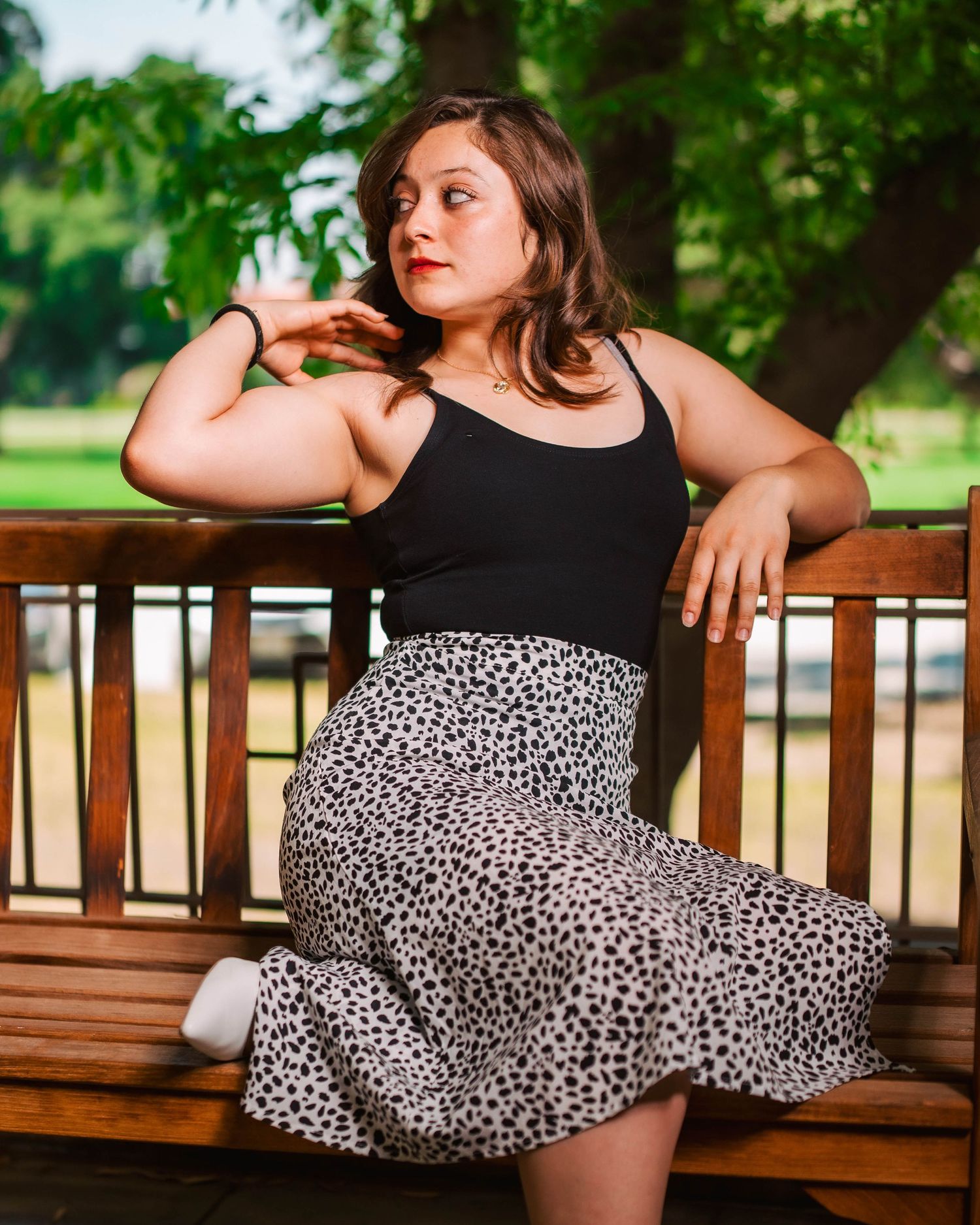 A person in a black tank top and leopard print skirt poses on a wooden bench in a park setting.