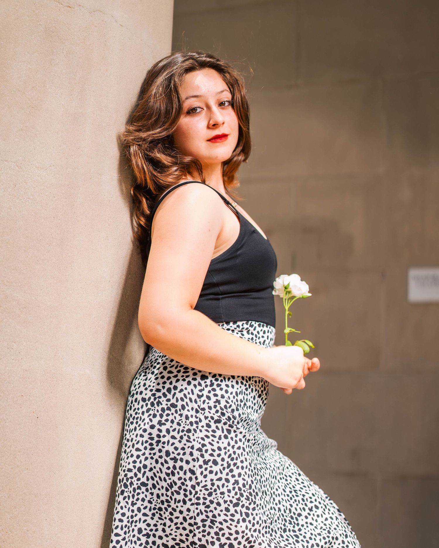 Person in black top and leopard print skirt poses against wall while holding white flower in natural lighting.