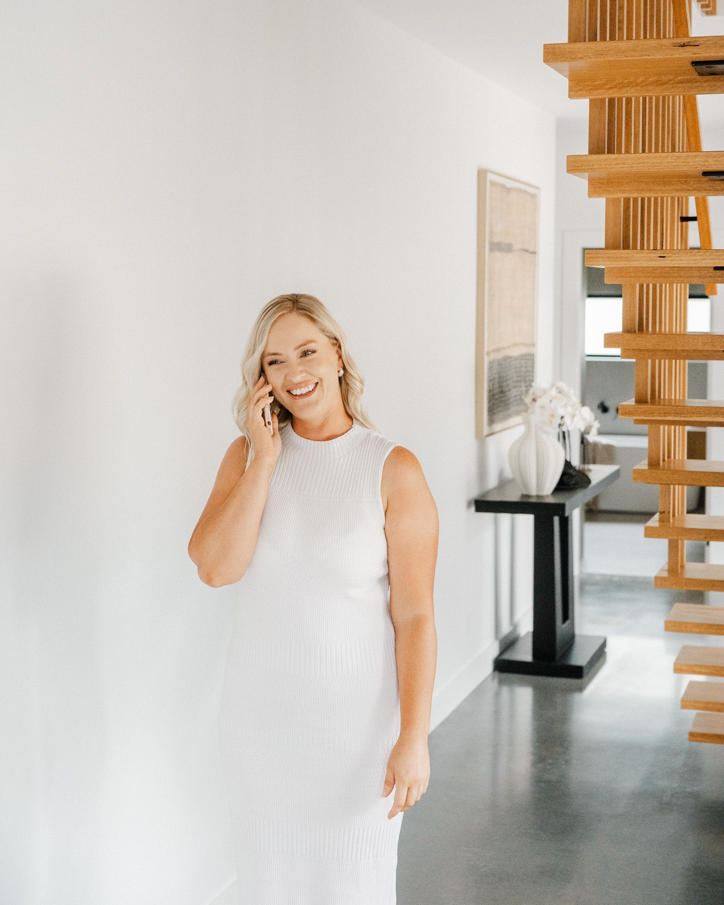A person in a white sleeveless top poses in a bright modern interior space with wooden beams and grey flooring.