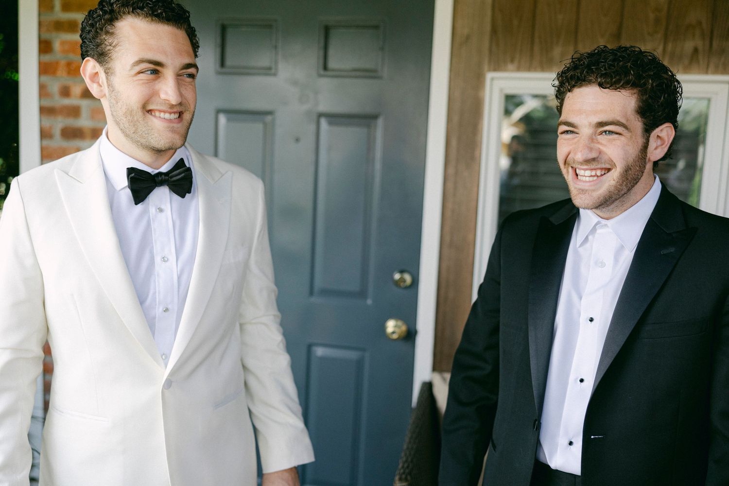 Two groomsmen in formal attire, one wearing a white tuxedo jacket and one in black, share a laugh before a wedding.