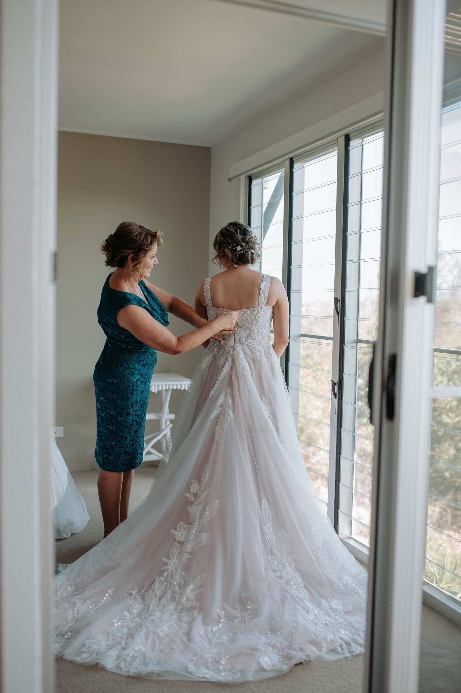 A woman in teal dress helps with a white lace wedding gown featuring an elegant long train by sunlit windows.