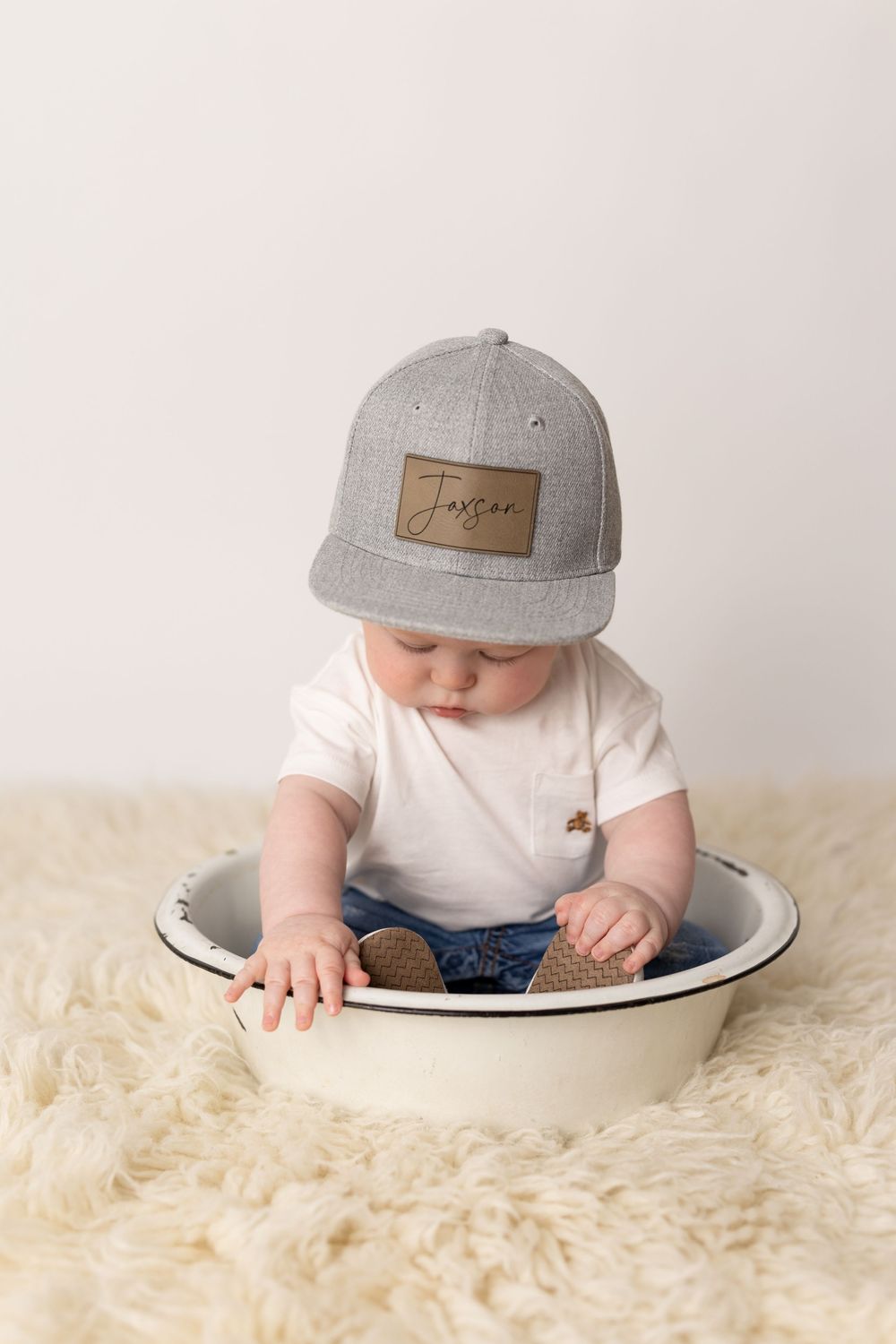 Baby in gray cap lies on white fluffy rug while reaching into ceramic bowl.