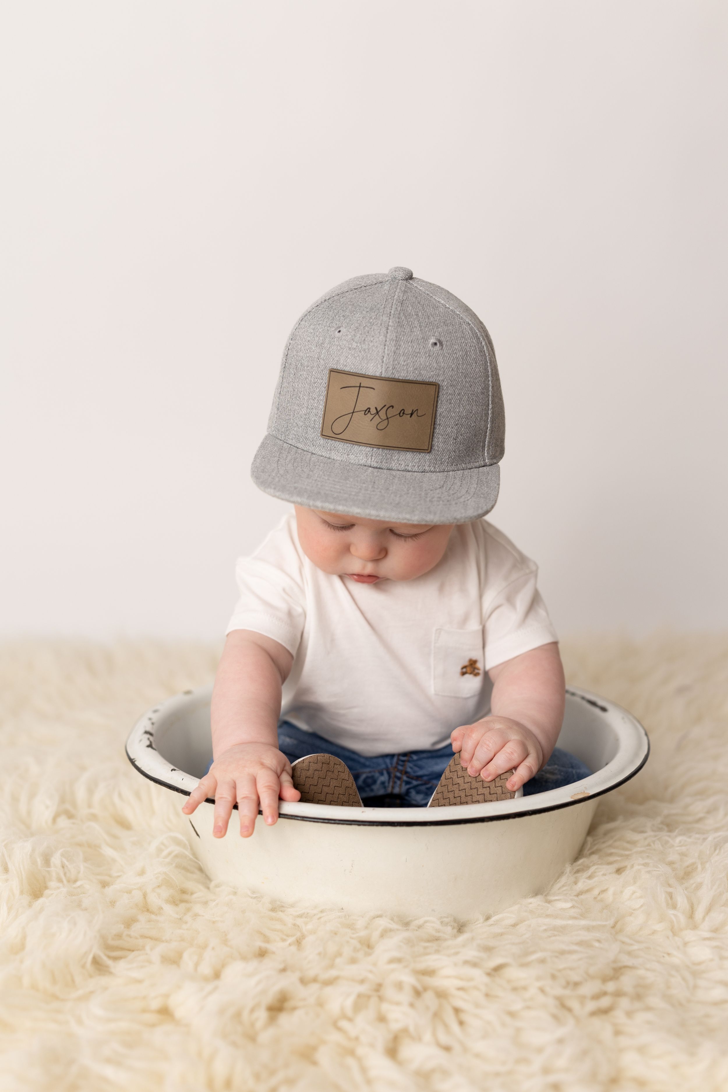 Baby in gray cap lies on white fluffy rug while reaching into ceramic bowl.