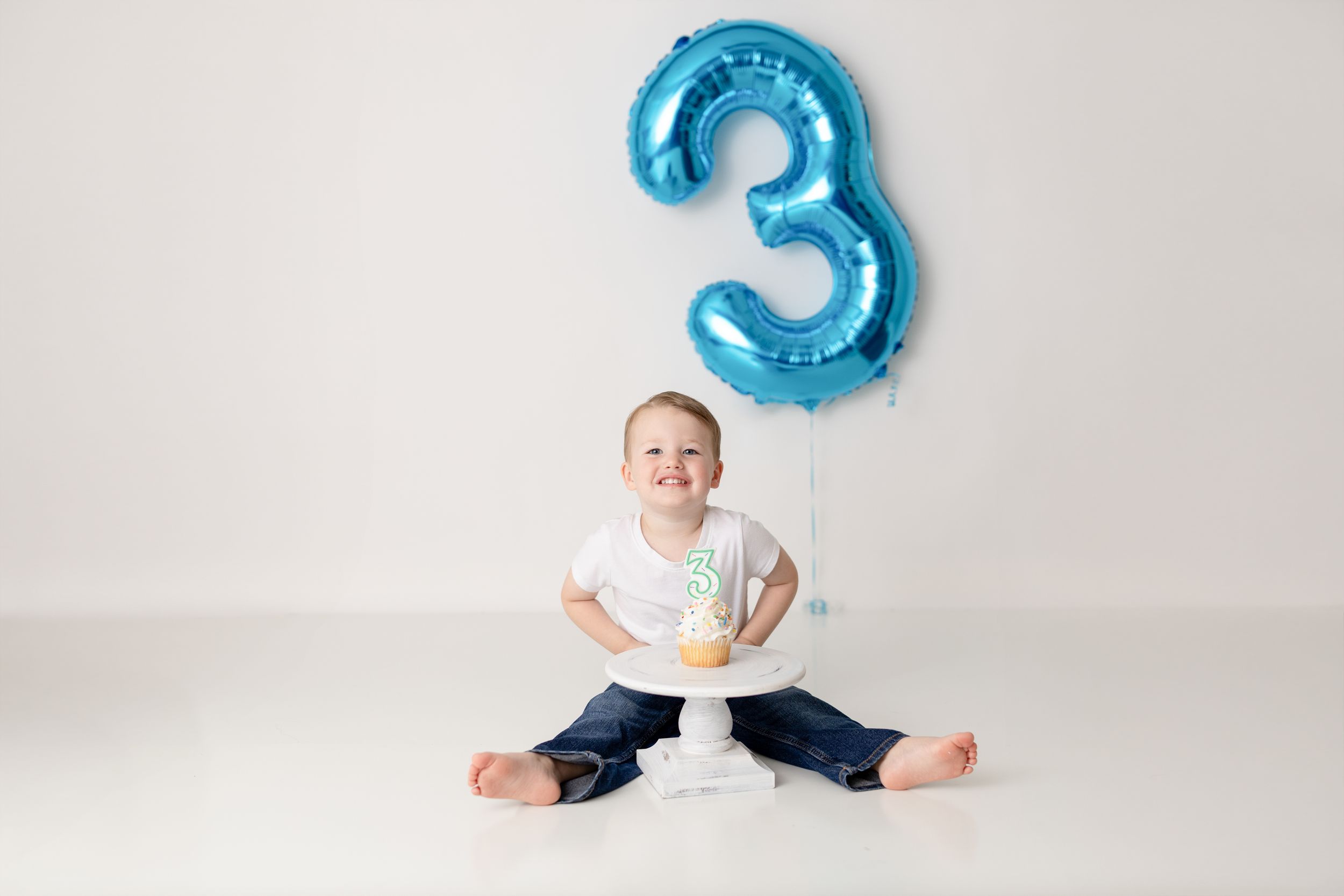 A child sits on a white backdrop with a large blue number 3 balloon for a birthday photo session.