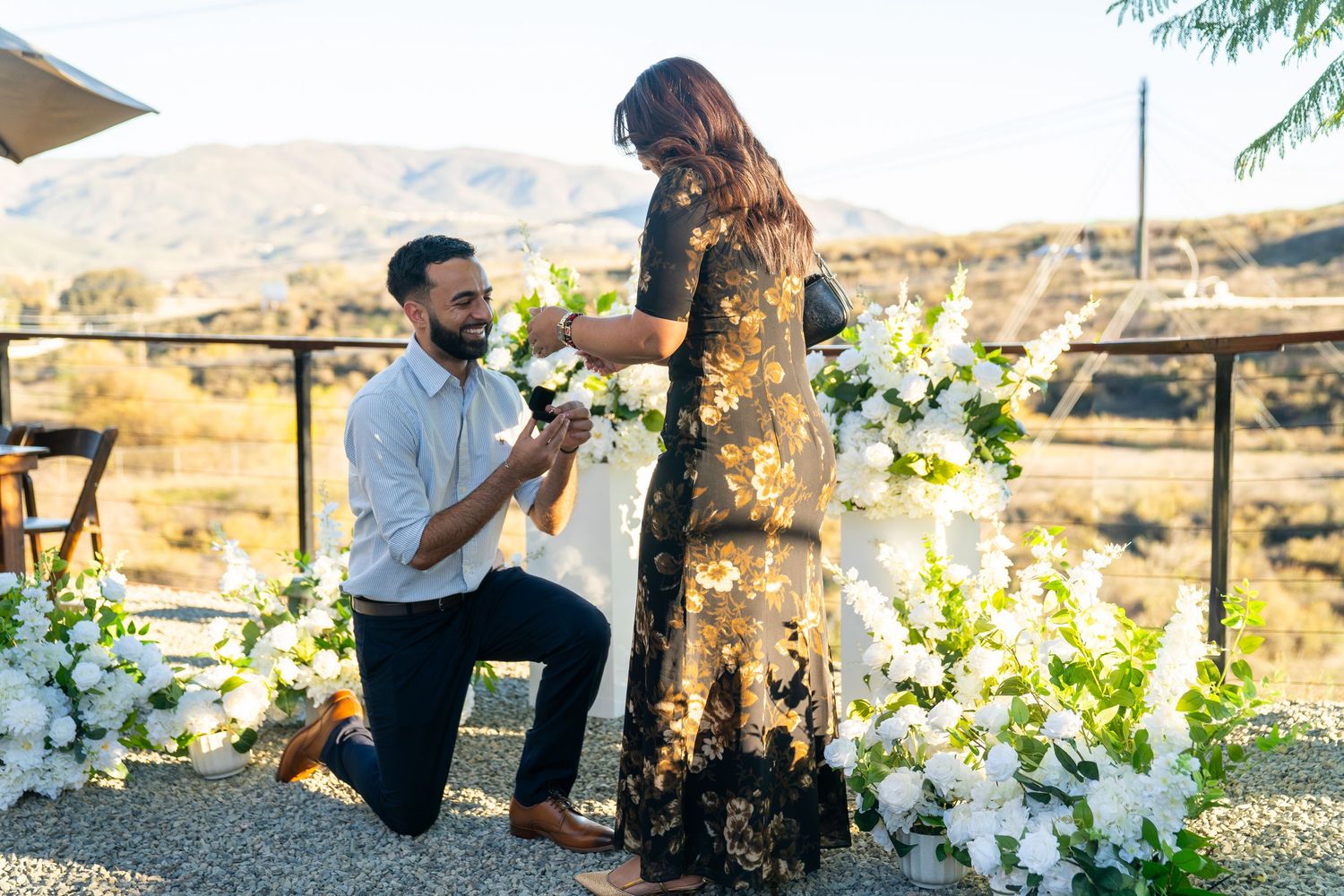 A romantic proposal moment outdoors with white floral decorations against a scenic mountain landscape background.
