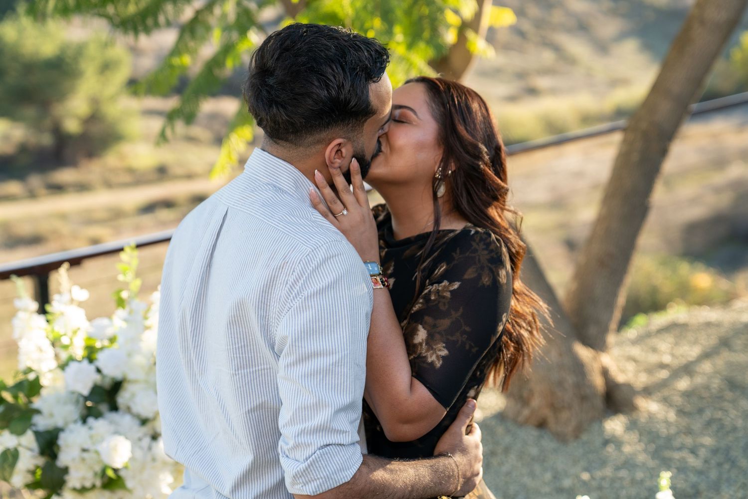 A couple shares romantic kisses in a sunlit outdoor setting with trees in the background.