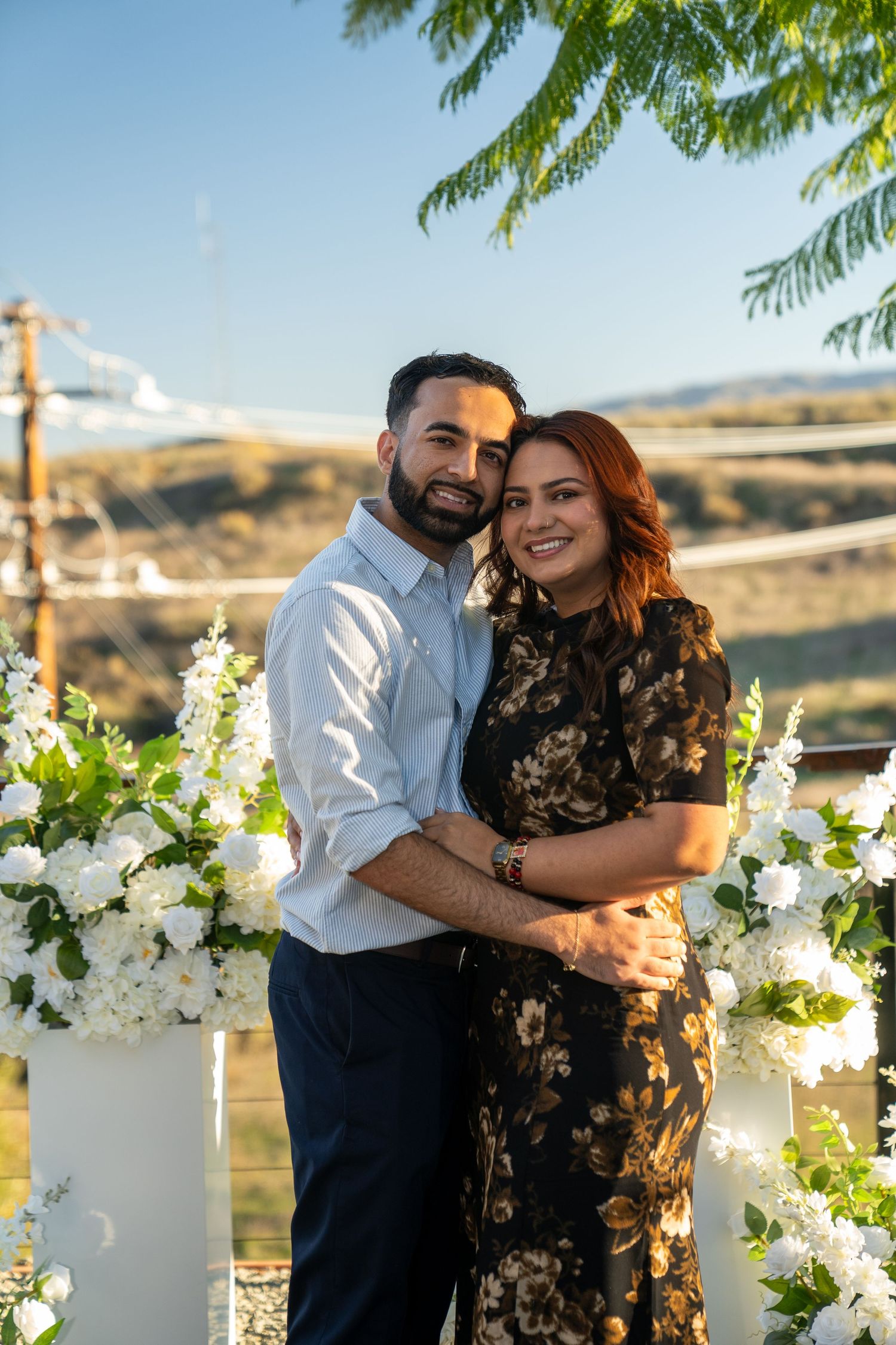 A couple embraces in front of white floral decorations with mountains in the background during a sunny outdoor event.