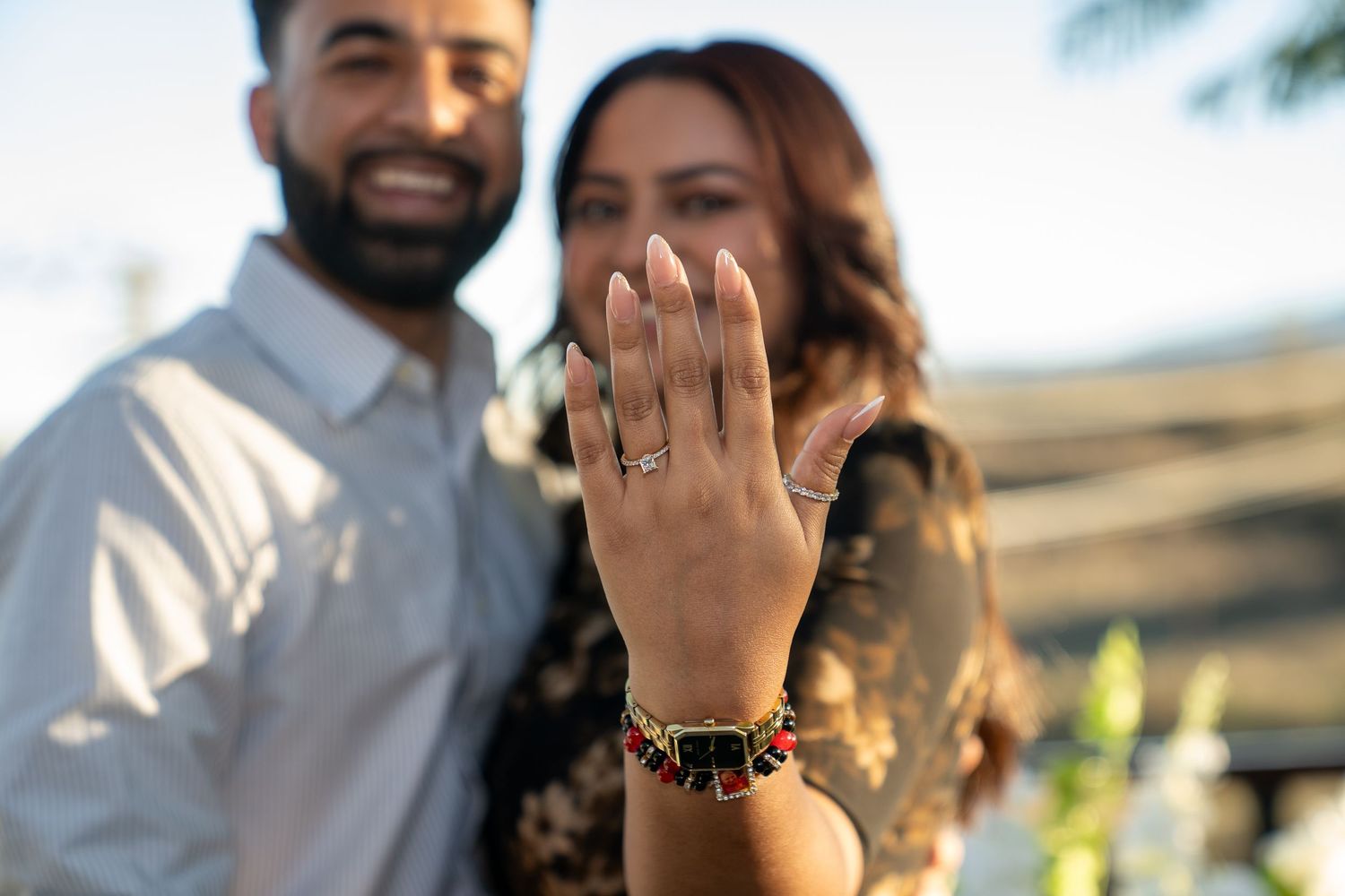 A hand wearing a new engagement ring is held up in focus while a couple stands blurred in the background.