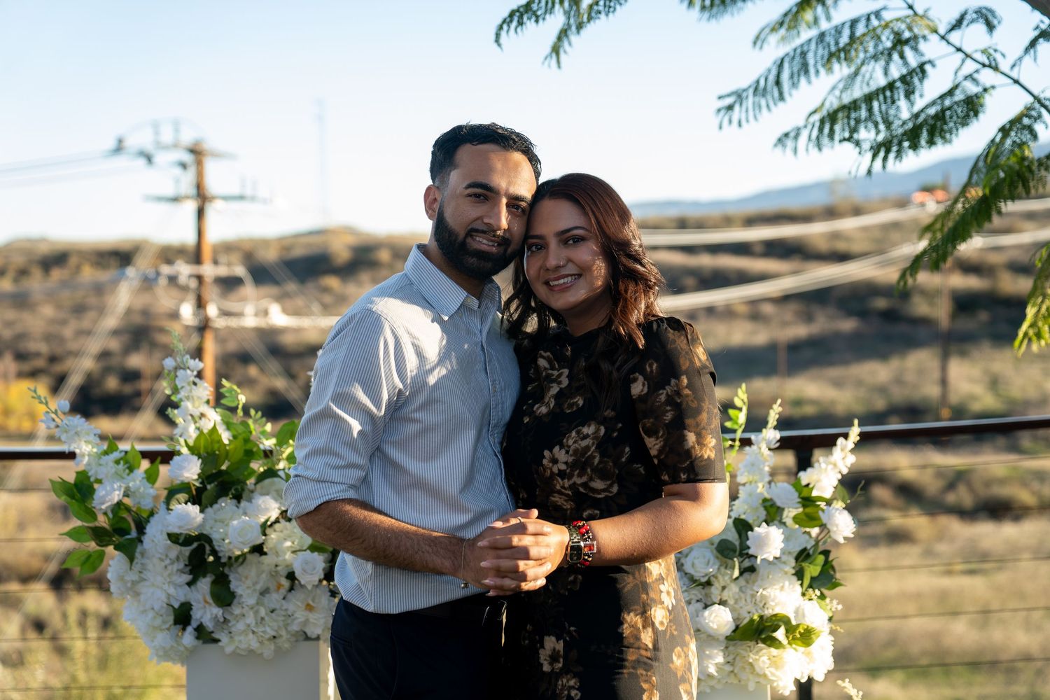 A couple embraces outdoors with white floral arrangements and rolling hills in the background.
