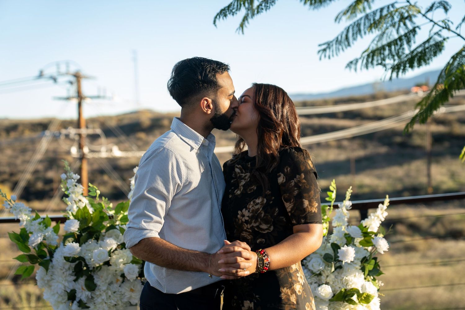 A couple embraces during a sunset photoshoot in front of palm trees and mountains in a tropical setting.