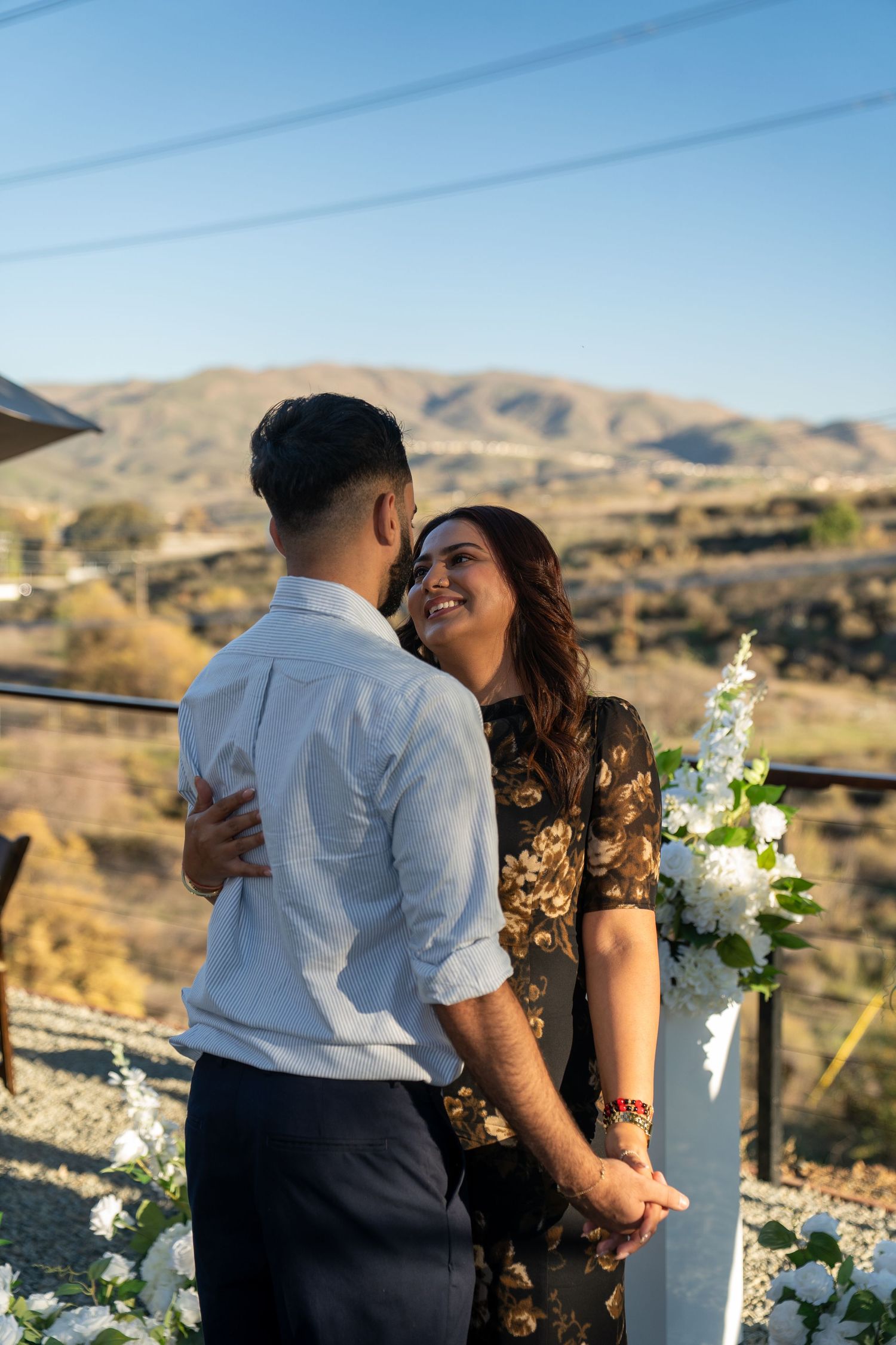A couple shares a romantic moment on a scenic outdoor deck with desert mountains and white flowers in the background.
